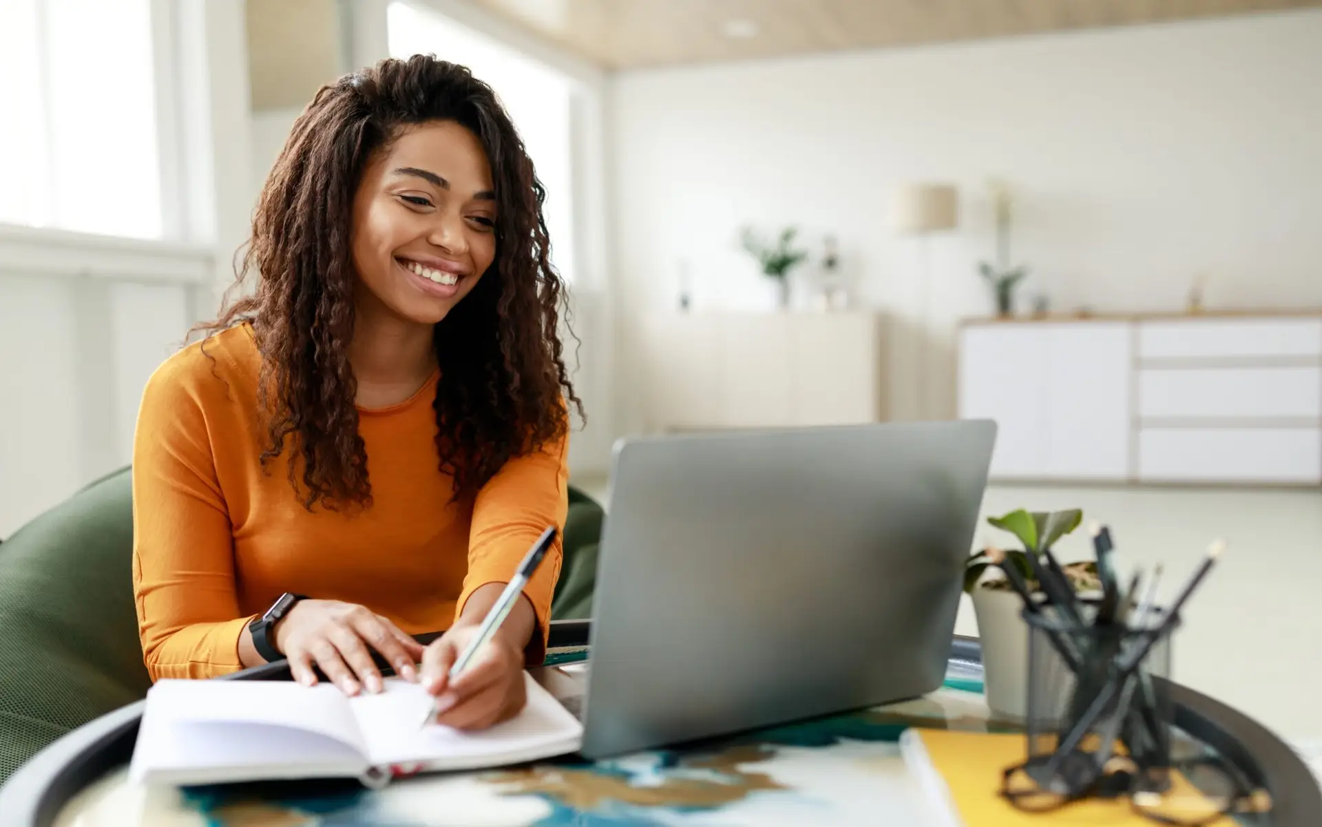 Young African American woman showing resilience while working in a hybrid environment, taking notes during an online meeting.