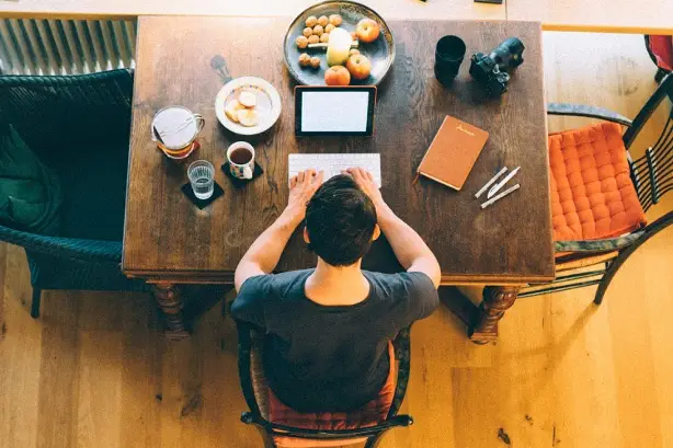 Man working from home at a dining table, typing on a tablet with coffee and fruit nearby, illustrating the importance of setting boundaries for healthy work-life balance