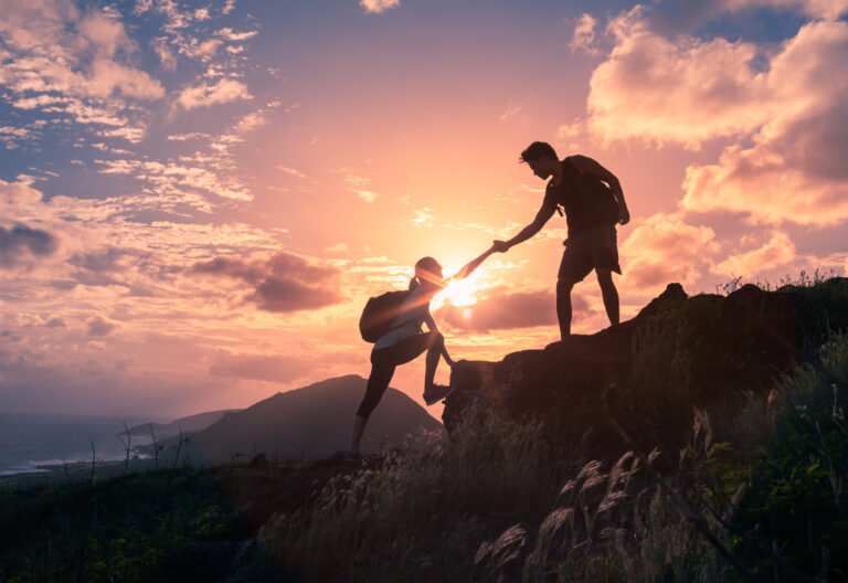 Male and female hikers showing resilience as they climb a mountain cliff, with one offering a helping hand during a hybrid outdoor adventure.
